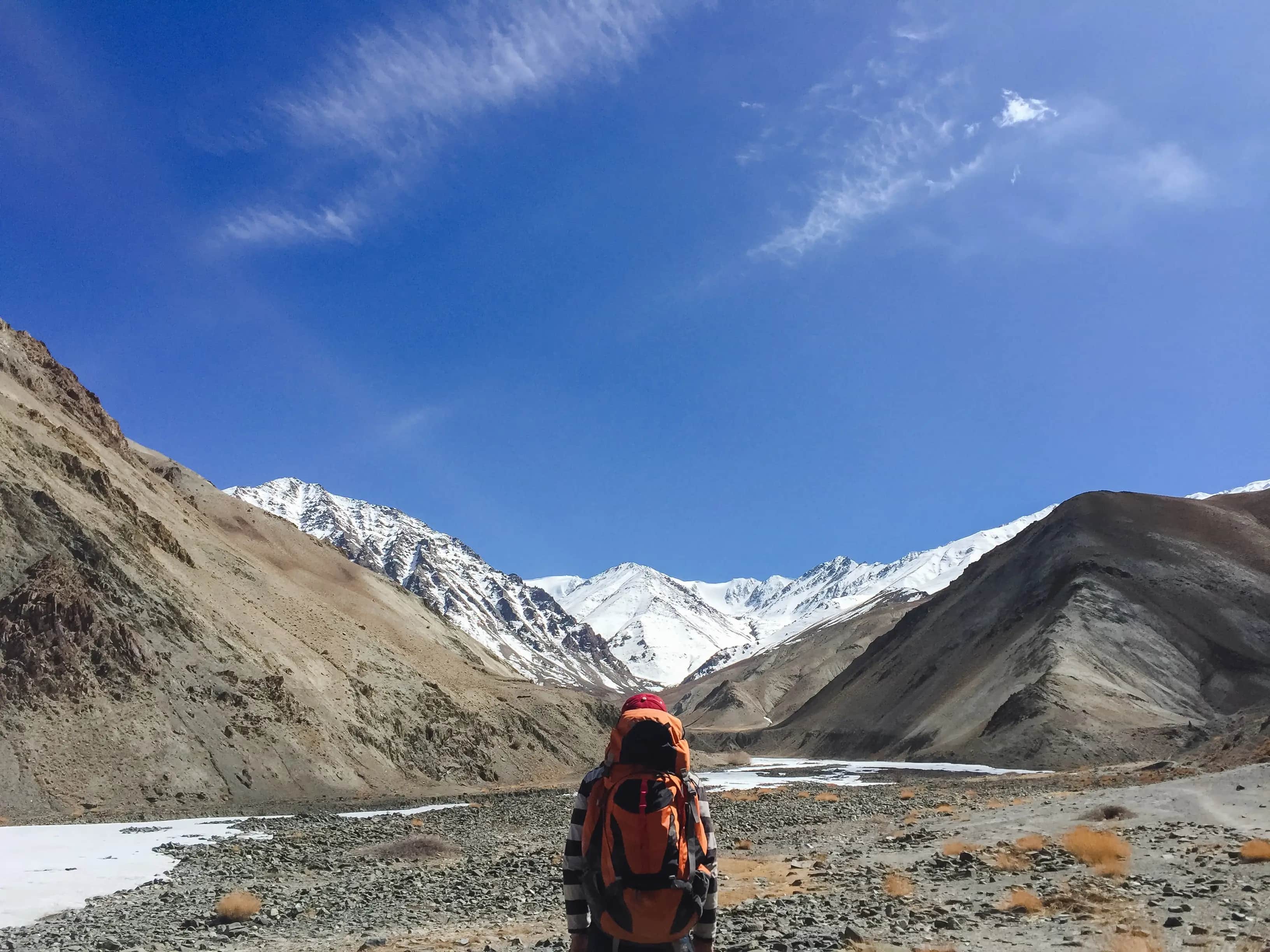 Image for A hiker with an orange backpack trekking through a rocky valley toward snow-capped mountains on the Upper Dolpo Trek in Nepal under a clear blue sky.