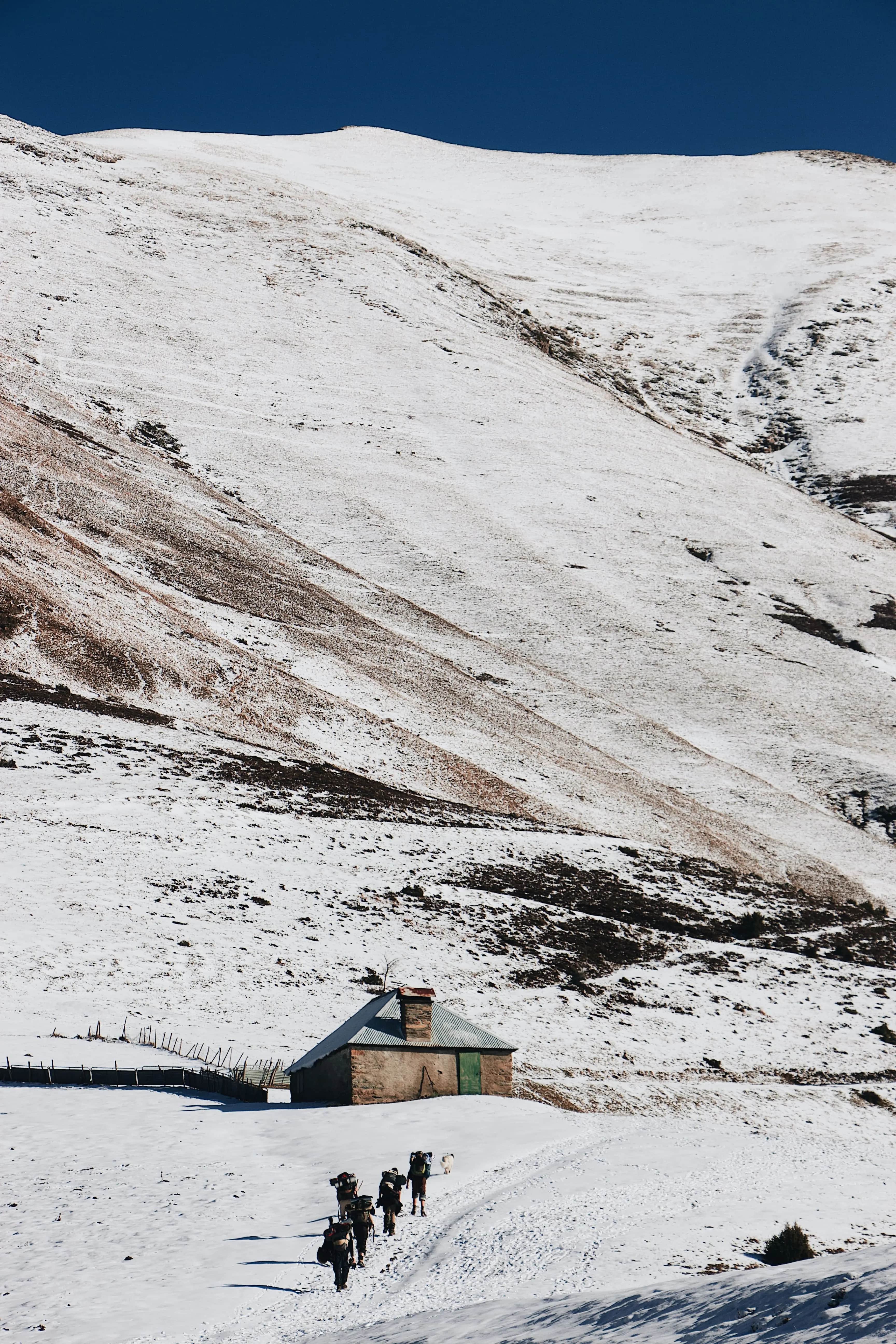 Image for High-angle view of hikers navigating a snowy trail in the remote Upper Mustang region of Nepal, approaching a rustic mountain shelter under a deep blue sky.